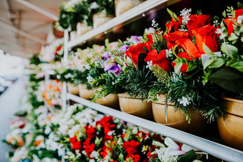 Two street vendors are seated outdoors at a flower stall, surrounded by an assortment of colorful fresh flowers arranged in bouquets and small vases. The flowers include roses, chrysanthemums, and oth