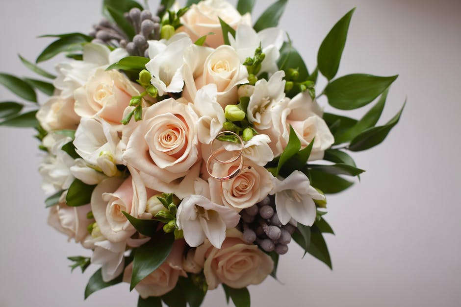 A bouquet of white flowers resting on a weathered wooden bench, including white calla lilies, roses, and small baby's breath, accented with soft green foliage. The bouquet is loosely arranged, showcas