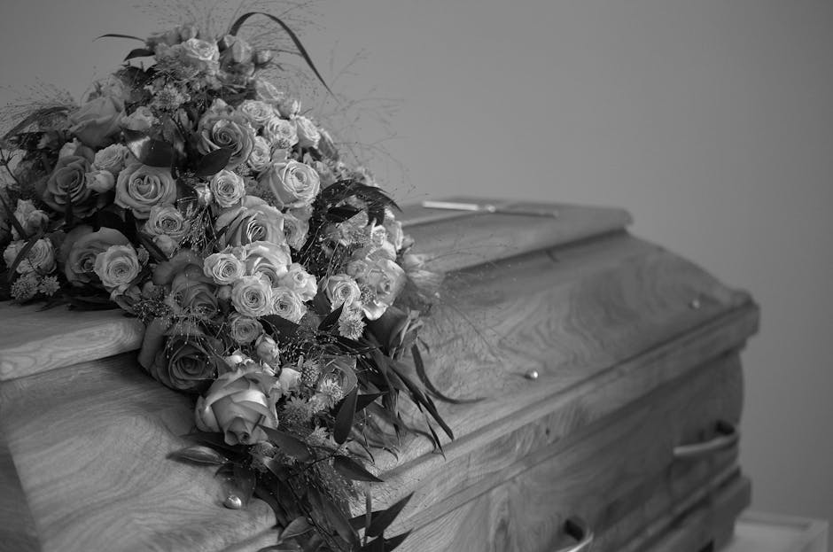 A group of six people sitting in a room with large windows, attending a funeral ceremony. The woman in the center, holding a bouquet of flowers, appears to be emotional. The setting features dark, ele